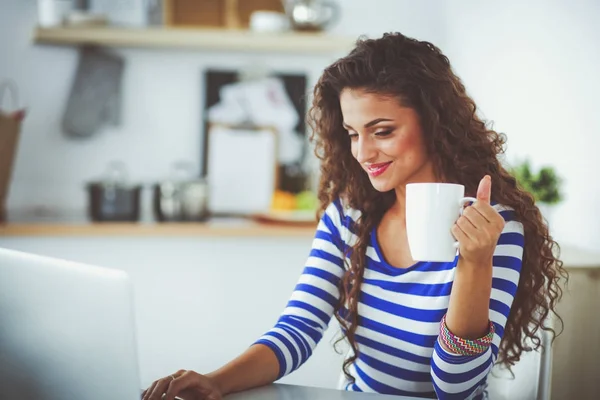 Lachende jonge vrouw met koffiekopje en laptop in de keuken thuis. Lachende jonge vrouw — Stockfoto