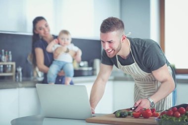 Masadaki dizüstü bilgisayara bakarken mutfakta sebze pişiren adamın portresi.