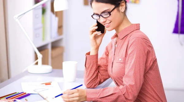 Giovane attraente stilista di moda femminile che lavora alla scrivania dell'ufficio, disegnando mentre parla sul cellulare — Foto Stock