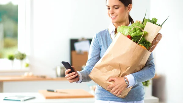 Mujer joven sosteniendo bolsa de la compra de comestibles con verduras. De pie en la cocina — Foto de Stock