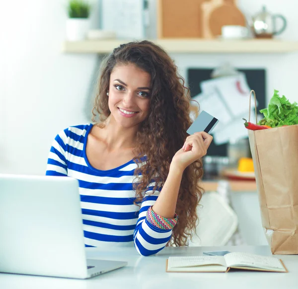 Mujer sonriente compras en línea utilizando la computadora y la tarjeta de crédito en la cocina —  Fotos de Stock