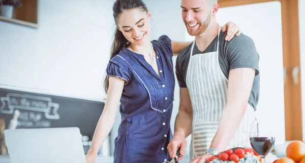 Junger Mann schneidet Gemüse und Frau steht mit Laptop in der Küche — Stockfoto