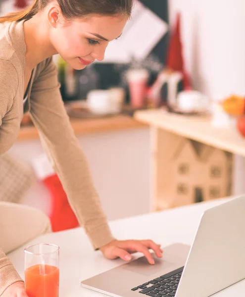 Mujer usando un portátil mientras bebe jugo en su cocina — Foto de Stock