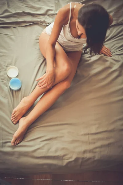 Wanita cantik duduk di tempat tidur dan menerapkan krim di kaki — Stok Foto