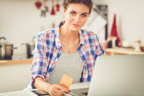 Mujer sonriente compras en línea utilizando la computadora y la tarjeta de crédito en la cocina. Mujer sonriente — Foto de Stock