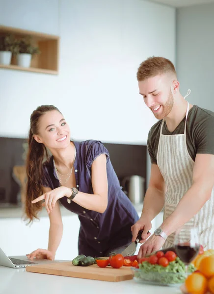 Hombre joven cortando verduras y mujer de pie con el ordenador portátil en la cocina — Foto de Stock