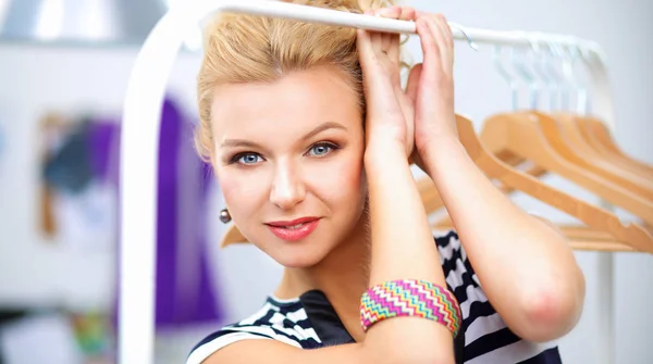 Beautiful young stylist near rack with hangers in office