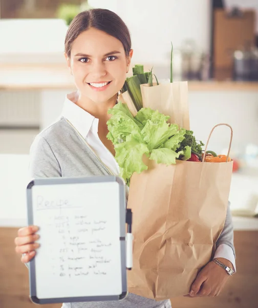 Mujer joven sosteniendo bolsa de la compra de comestibles con verduras. De pie en la cocina —  Fotos de Stock