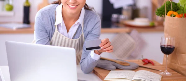 Mujer sonriente compras en línea utilizando la computadora y la tarjeta de crédito en la cocina —  Fotos de Stock