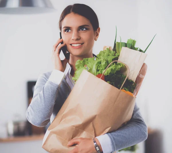 Mujer sonriente con teléfono móvil sosteniendo bolsa de compras en la cocina —  Fotos de Stock