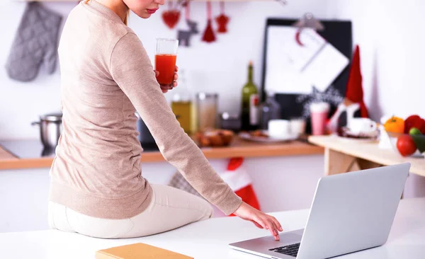 Mulher usando um laptop enquanto bebe suco em sua cozinha — Fotografia de Stock