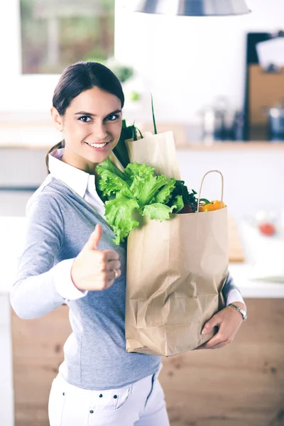 Mujer joven sosteniendo bolsa de la compra de comestibles con verduras. De pie en la cocina — Foto de Stock