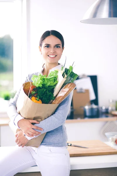 Mujer joven sosteniendo bolsa de la compra de comestibles con verduras. De pie en la cocina —  Fotos de Stock