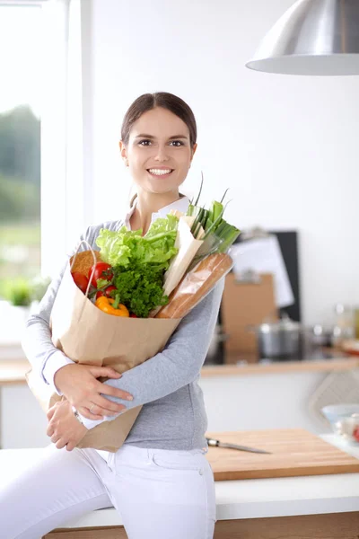 Mujer joven sosteniendo bolsa de la compra de comestibles con verduras. De pie en la cocina — Foto de Stock