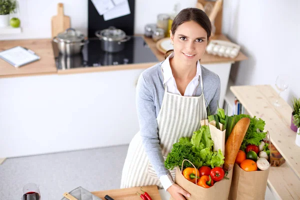 Mujer joven sosteniendo bolsa de la compra de comestibles con verduras. De pie en la cocina —  Fotos de Stock