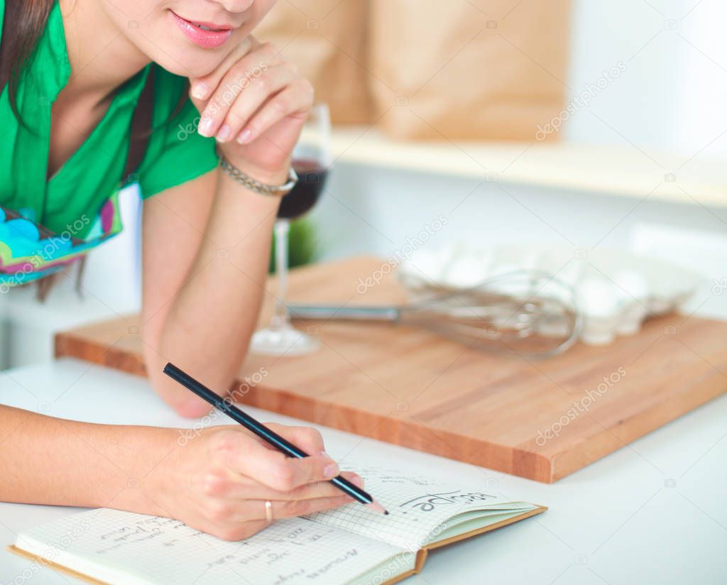 Happy beautiful woman standing in her kitchen writing on a notebook at home