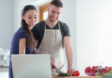 Sebze doğrayan genç bir adam ve mutfakta dizüstü bilgisayarı olan bir kadın.