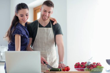 Sebze doğrayan genç bir adam ve mutfakta dizüstü bilgisayarı olan bir kadın.