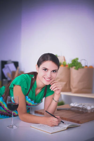 Счастливая красивая женщина, стоящая на кухне и пишущая на блокноте дома — стоковое фото