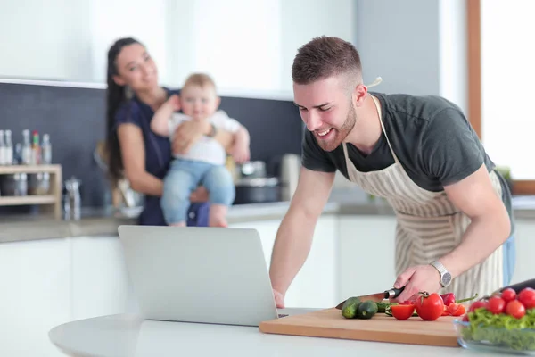 Retrato del hombre cocinando verduras en la cocina mientras mira una computadora portátil en la mesa — Foto de Stock