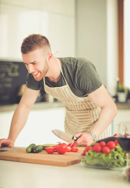 Retrato del hombre cocinando verduras en la cocina mientras mira una computadora portátil en la mesa — Foto de Stock