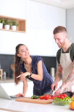 Sebze doğrayan genç bir adam ve mutfakta dizüstü bilgisayarı olan bir kadın.