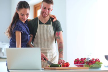 Sebze doğrayan genç bir adam ve mutfakta dizüstü bilgisayarı olan bir kadın.
