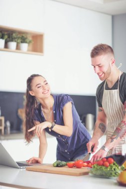 Sebze doğrayan genç bir adam ve mutfakta dizüstü bilgisayarı olan bir kadın.