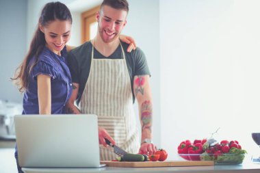 Sebze doğrayan genç bir adam ve mutfakta dizüstü bilgisayarı olan bir kadın.