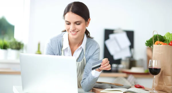 Senyum wanita belanja online menggunakan komputer dan kartu kredit di dapur — Stok Foto