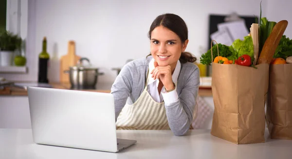 Mooie jonge vrouw koken op zoek naar laptop scherm met ontvangst in de keuken — Stockfoto