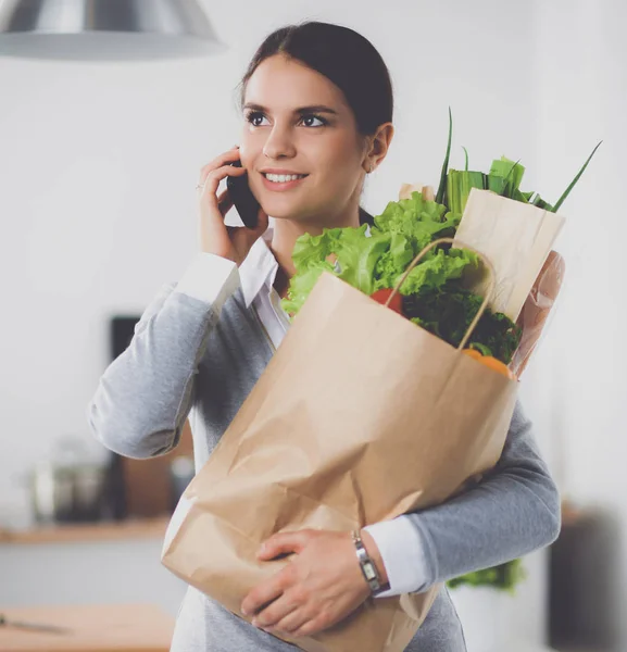 Leende kvinna med mobiltelefon håller shoppingväska i köket — Stockfoto