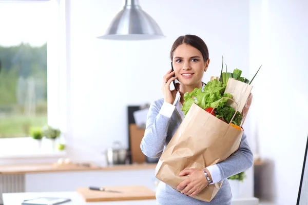 Mujer joven sosteniendo bolsa de la compra de comestibles con verduras. De pie en la cocina — Foto de Stock