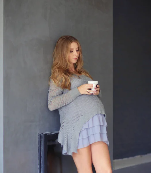 Беременная женщина с чашкой чая пьет чай, глядя в окно дома — стоковое фото