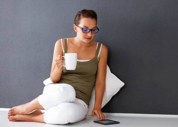 Krásná mladá žena s šálkem čaje, zatímco sedí na podlaze doma pomocí tabletového počítače. — Stock fotografie