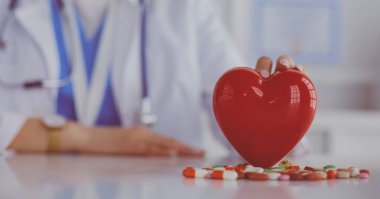 Beyaz önlüklü doktor. Steteskoplu. Elinde haplar ve beyaz bir kalbe takılmış plastik kalp var.