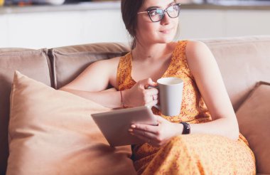 Genç kadın evinde kanepede kahve içerken tablet kullanıyor.