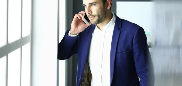 Homem de negócios de terno falando ao telefone e olhando para longe perto da janela . — Fotografia de Stock