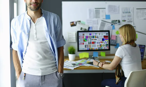 Porträt eines jungen Designers vor Laptop und Computer während der Arbeit. Assistentin bedient ihr Handy im Hintergrund. — Stockfoto