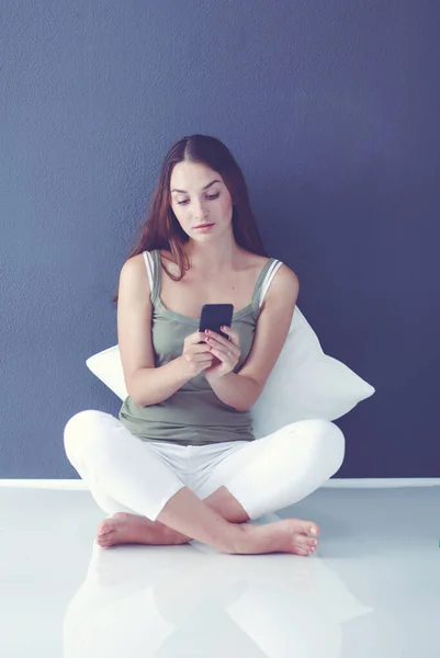 Souriant belle brune assise sur le sol et utilisant son téléphone dans le salon — Photo