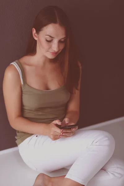 Mujer sentada en el suelo y mensajería de texto en un teléfono móvil aislado sobre fondo gris — Foto de Stock