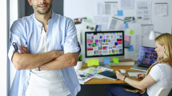 Porträt eines jungen Designers vor Laptop und Computer während der Arbeit. Assistentin bedient ihr Handy im Hintergrund. — Stockfoto