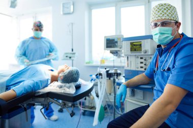 Ameliyat odasında arka planda erkek cerrah var.