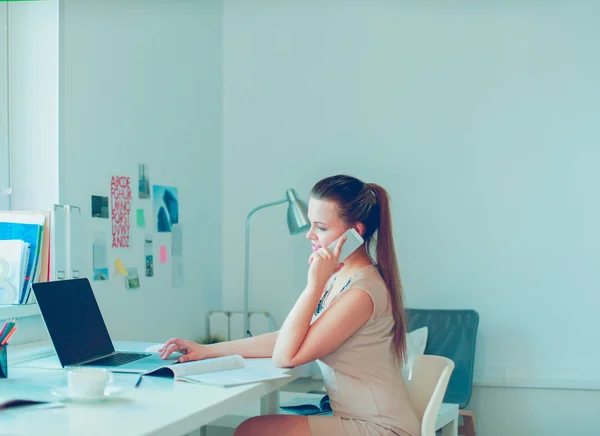 Attractive woman sitting at desk in office, working with laptop computer, holding document — Stock Photo, Image