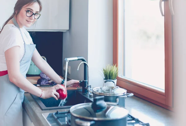 Nő zöldséget mos. Gyönyörű fiatal nő mosás zöldség saláta és mosolygós, miközben áll a konyhában — Stock Fotó