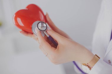 Steteskoplu bir doktor, beyaz arka planda kırmızı kalbi inceliyor.