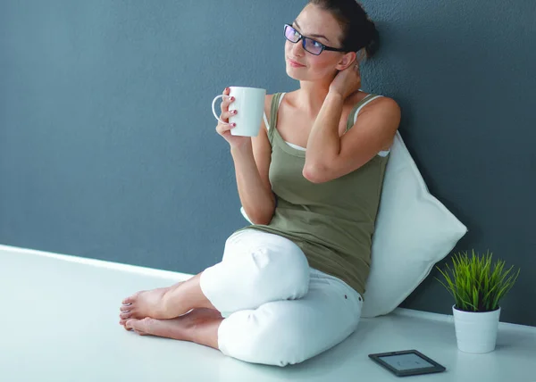 Μια όμορφη νεαρή υγιή κάθεται στο πάτωμα με ένα φλιτζάνι τσάι ή καφέ χαμογελώντας. απομονωμένο σε λευκό φόντο — Φωτογραφία Αρχείου