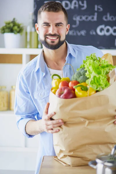 Man met papieren zak vol boodschappen op de keukenachtergrond. Winkelen en gezond voedsel concept — Stockfoto