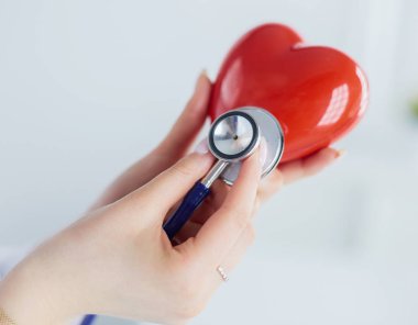 Steteskoplu bir doktor, beyaz arka planda kırmızı kalbi inceliyor.