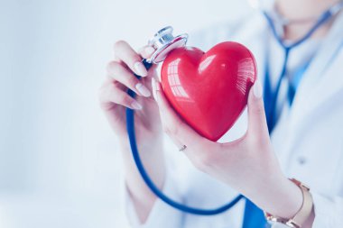Steteskoplu bir doktor, beyaz arka planda kırmızı kalbi inceliyor.